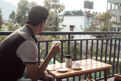 Mature adult tourist Solo business traveller of Indian ethnicity in casual clothing sitting on balcony of a holiday villa and holding a hot cup of tea in morning on Mountain city background. Travel vacation active life and healthy lifestyles concept.