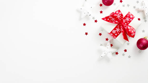 istock Top view Christmas present and festive ornaments on white background. Flat lay gift box with red ribbon bow, white decorations, red balls, confetti stars. Xmas greeting card with copy space 1185621407