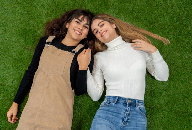 Top view of two stylish girl friends having fun laughing and making faces lying down on grass. Blonde and brunette happy young teenagers women enjoying time together. Friendship, love and care.
