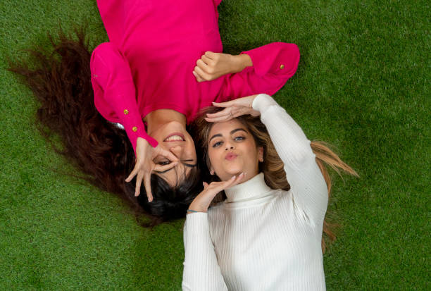 Top view of two stylish girl friends having fun laughing and making faces lying down on grass. Blonde and brunette happy young teenagers women enjoying time together. Friendship, love and care.