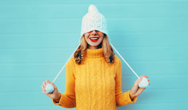 istock Winter portrait happy smiling young woman having fun pulls a hat over her eyes wearing yellow knitted sweater and white hat with pom pom on blue wall background 1185648851