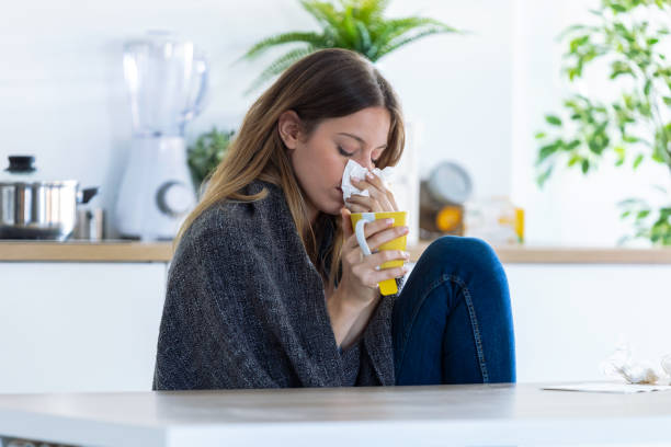 Illness young woman sneezing in a tissue while drinking hot beverage in the kitchen at home.