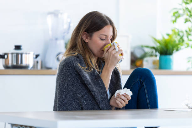 istock Sick and cold young woman drinking hot beverage while sitting in the kitchen at home. 1185653017
