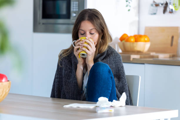 istock Sick and cold young woman drinking hot beverage while sitting in the kitchen at home. 1185653065