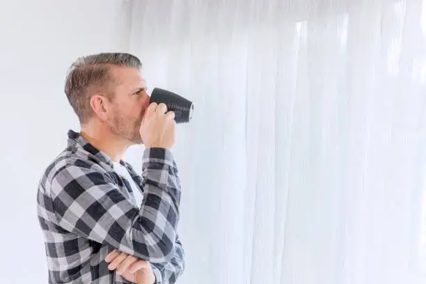 Side view of Caucasian man drinking a cup of coffee while looking out the window at home