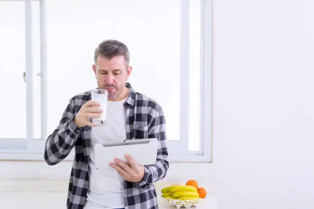 Caucasian man drinking a glass of milk while using a digital tablet and standing near the window at home