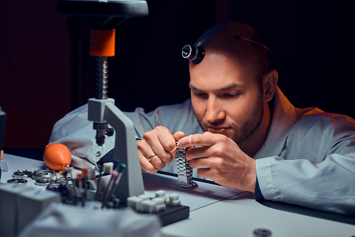 Expirienced watchmaker is doing engraving for custmer's watch at his workshop.