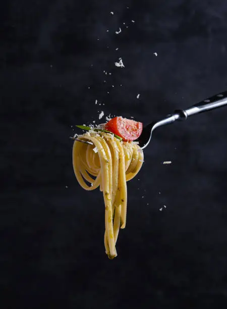 Italian tagliatelle with tomato, sause pesto, basil and flying parmesan on black fork. Image minimalism and dark background.