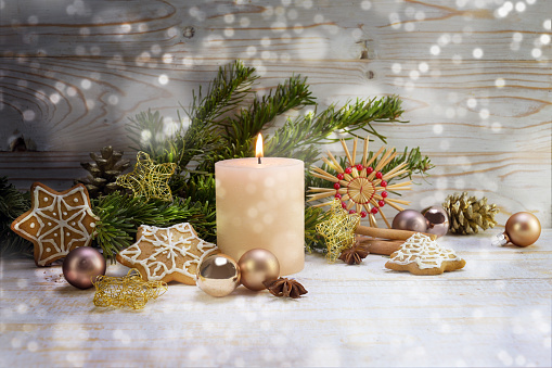 Burning candle with Christmas decoration, fir branches, baubles, straw stars and gingerbread on rustic wood, snowy boheh lights, copy space, selected focus