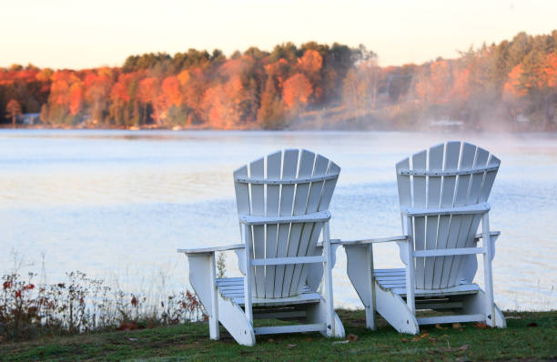 A row of colourful adirondack chairs on a dock in fall. A boat is also in the scene beside the dock. Tranquil morning with no wind. Nobody is in the image. Fall at the cottage theme.