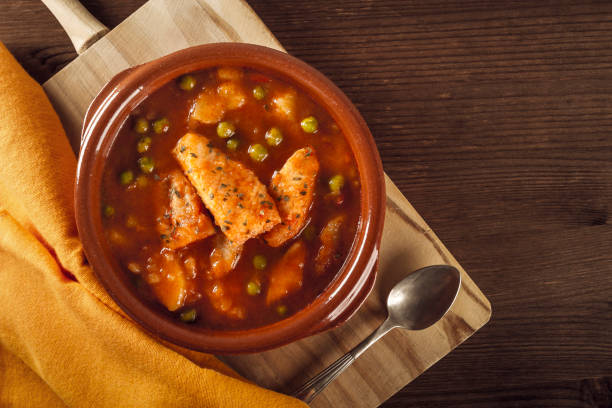 Top view of a clay pot with a delicious fish stew with peas and cooked potatoes with a vintage spoon and an orange napkin on a rustic table. Healthy fish soup. Homemade food. Hot food, freshly made stock photo