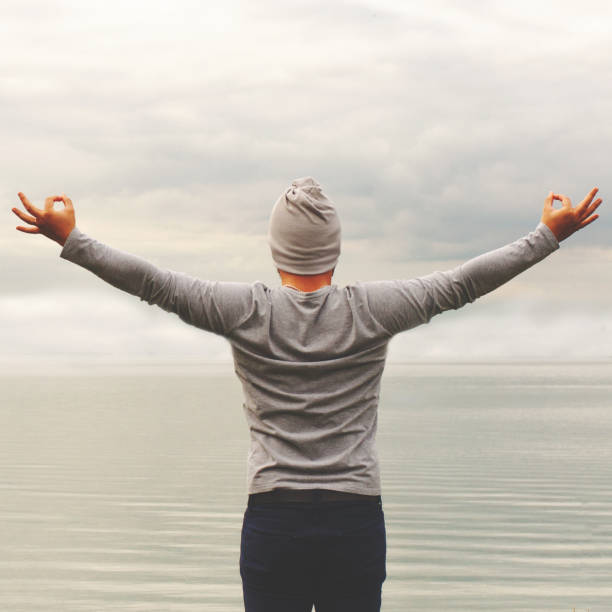 istock A young man is standing on the shore. The view from the back. Yoga classes. Hands raised up. Shows with hands OK sign. Freedom and achievement. 1185760842