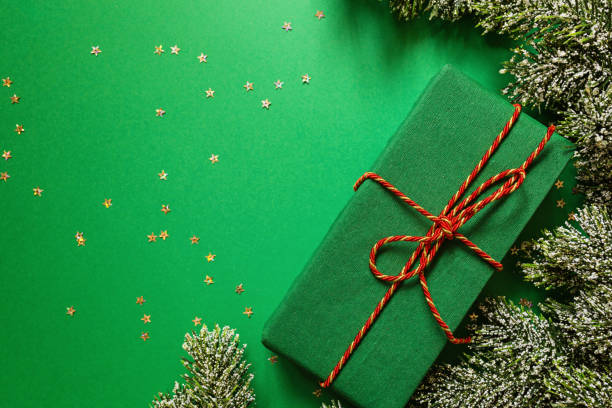 istock christmas wrapped gift box and tree branches on green background with confetti. new year concept. Greeting card, xmas celebration 2020. Flat lay, template, top view, copy space 1185769081
