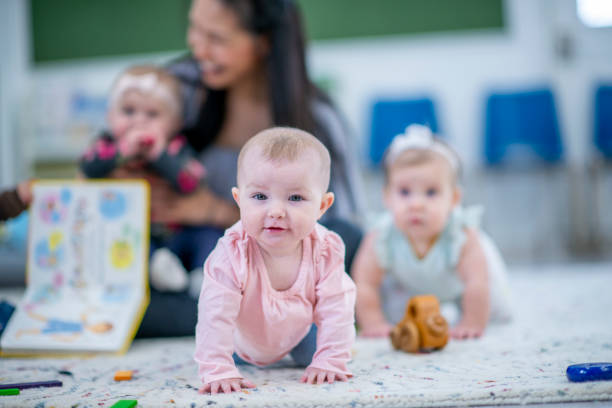 Little Girl Crawling in a Daycare Room stock photo A little Caucasian girl crawls among other multi-ethnic babies on a daycare room floor. She is smiling and enjoying her pay time. A daycare worker is sitting in the background with another little girl on her lap. There are toys and storybooks out around the room. asian nanny stock pictures, royalty-free photos & images