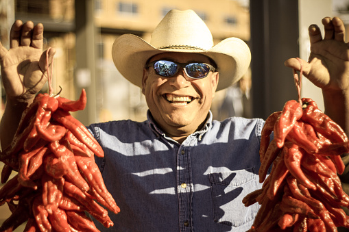 Santa Fe, NM: A laughing farmer holding up two chili pepper ristras for sale at the Saturday Farmers Market located in the Santa Fe Railyard.