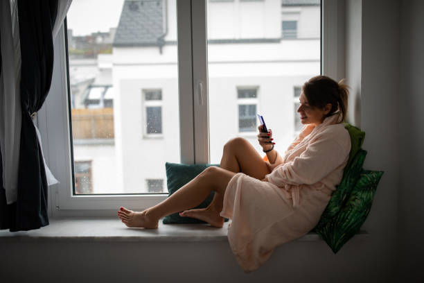 Beautiful young woman at home, sitting on window sill and using phone
