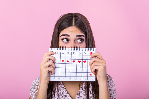 Retrato de una joven divertida escondida detrás de un calendario de períodos menstruales y mirando hacia otro lado en el espacio de copia aislado sobre fondo rosa. Calendario del Período Femenino photo