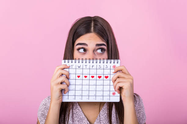 retrato de una joven divertida escondida detrás de un calendario de períodos menstruales y mirando hacia otro lado en el espacio de copia aislado sobre fondo rosa. calendario del período femenino - menstruación fotografías e imágenes de stock