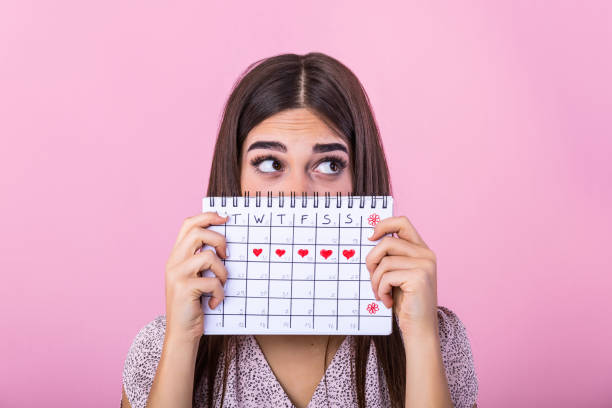 Portrait of a funny young girl in hiding behind a menstrual periods calendar and looking away at copy space isolated over pink background. Female Period calendar Portrait of a funny young girl in hiding behind a menstrual periods calendar and looking away at copy space isolated over pink background. Female Period calendar menstruation photos stock pictures, royalty-free photos & images