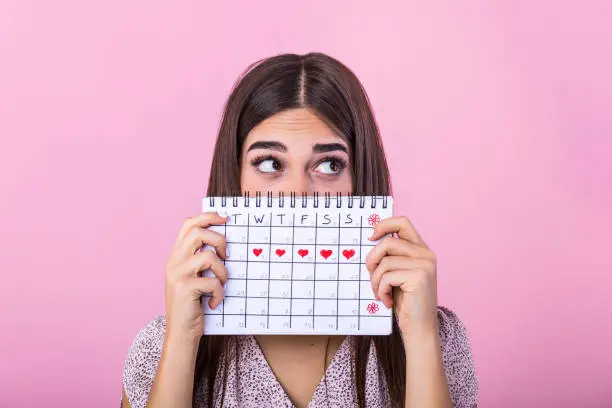 Photo of Portrait of a funny young girl in hiding behind a menstrual periods calendar and looking away at copy space isolated over pink background. Female Period calendar