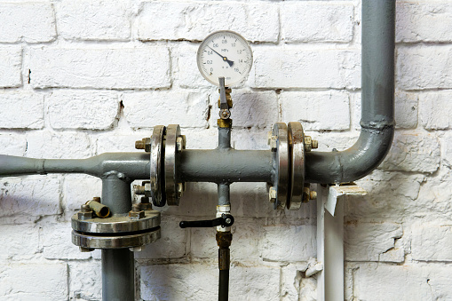 heating system pipe section with pressure gauge, valve and fittings, against a plastered brick wall