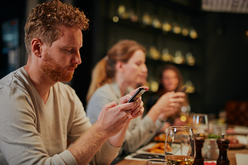 Small group of friends sitting in restaurant for dinner and using smartphones.