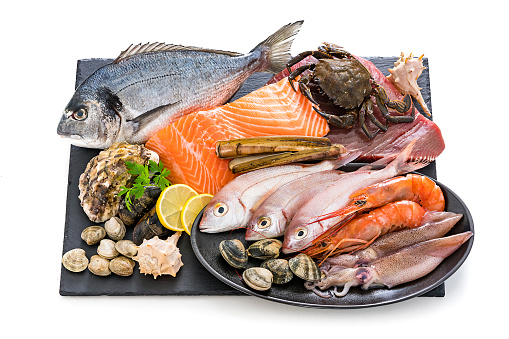 Food: group of healthy fresh raw seafood shot on white background. The composition is arranged on a black slate board and includes fish, salmon steak, tuna steak, crab, shrimps, razor clams, mussels, oyster, squid and various clams. Predominant colors are red, brown and white. XXXL 42Mp studio photo taken with Sony A7rii and Sony FE 90mm f2.8 macro G OSS lens