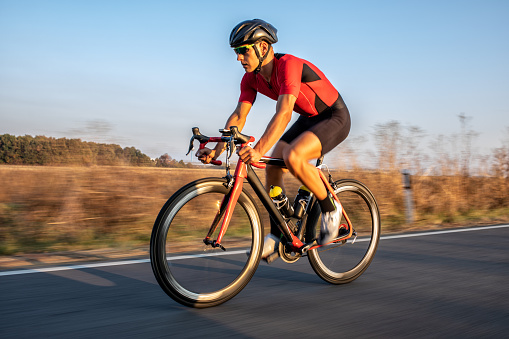 Young professional athlete riding bicycle for exercise on the road through the countryside. He is well equipped, with protective helmet, sunglasses, black and red jersey. Photo taken when cyclist is in motion, from car in motion, for speed effect.