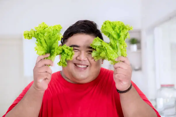 Close up of an obese man smiling at the camera while holding fresh lettuce in the kitchen