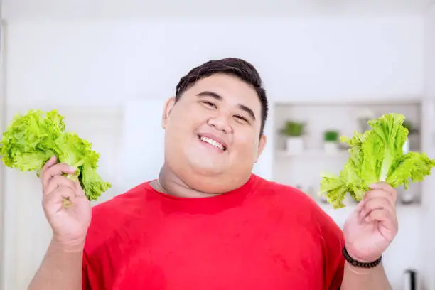 Picture of an overweight man smiling at the camera while holding fresh lettuce in the kitchen