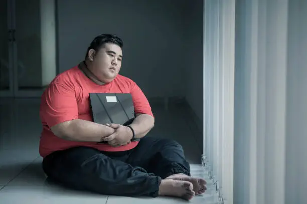 Picture of an obese man looks pensive while holding a weight scale and sitting near the window