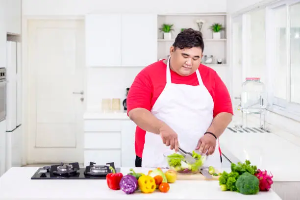Picture of young fat man mixing a bowl of fresh salad while standing in the kitchen