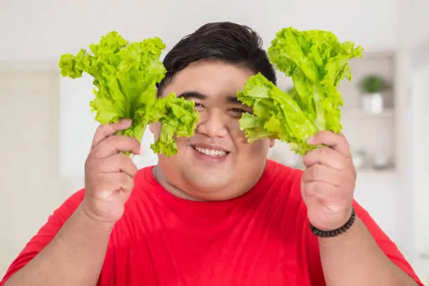 Close up of overweight man smiling at the camera while holding fresh lettuce in the kitchen