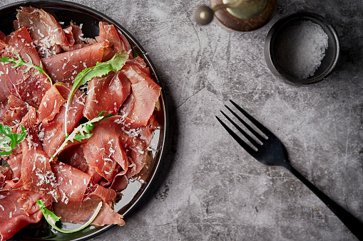 Tasty appetizing classic Italian bresaola with rocket salad and parmesan on black plate on black background. View form above. Top view. Horizontal. With copy space for text.