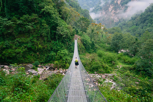 A rear-view shot of a caucasian male tourist standing next to a suspension bridge mid way through a Trek to Annapurna Base Camp in Nepal.