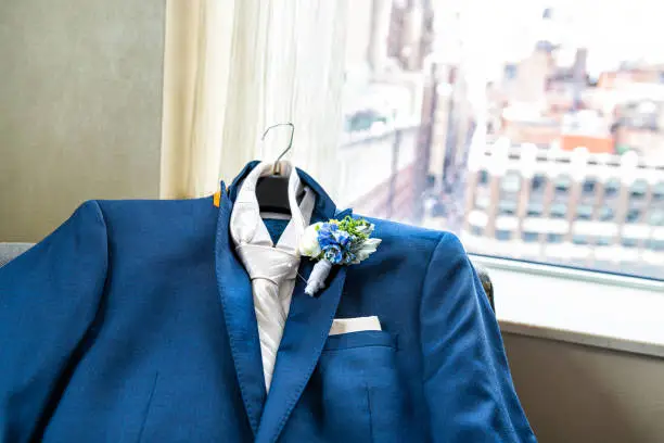 Men's suit for groom closeup with flower boutonniere, pin wedding preparation and pocket handkerchief by window view of urban New York City NYC