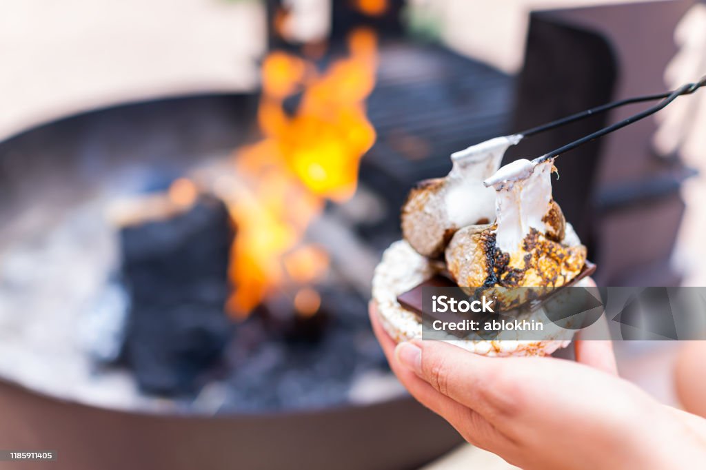 Young woman hand holding gooey roasted charred marshmallows smores with chocolate and rice cake cracker by fire in campground campfire grill Smore Stock Photo