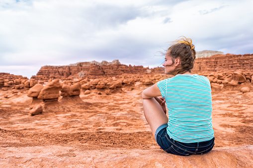 Woman hiker sitting back looking at hoodoo sandstone rock formations with cloudy storm sky and wind desert landscape in Goblin Valley State Park in Utah on trail