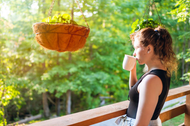 appendere piante in vaso in estate con donna in piedi sotto il portico di casa nel cottage della cabina di legno mattutino bevendo tè o caffè - balcony women patio coffee foto e immagini stock