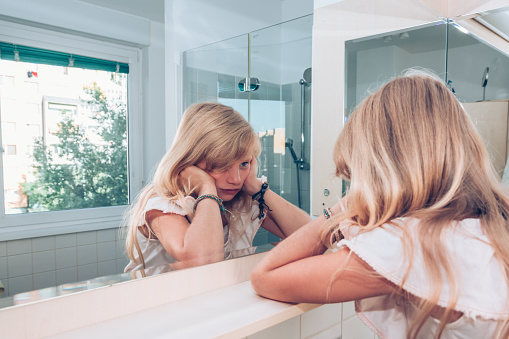 close up portrait of adorable girl with long blond hair in the bathroom with sad expression in the face while looking into mirror