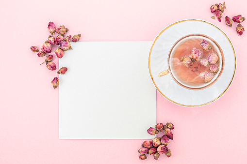 Overhead studio image of an elegant  steaming tea cup with made of Chinese dried roses. The background is pale pink. There is a blank card by the cup, and it is framed by dried rose buds. \n\n(The image coloring is based on PANTONE Blossom color nuances)