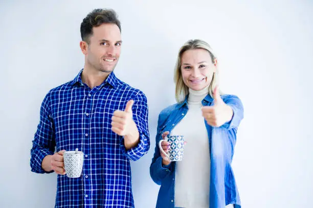 Photo of Young beautiful couple drinking cup of coffee standing over isolated white background happy with big smile doing ok sign, thumb up with fingers, excellent sign