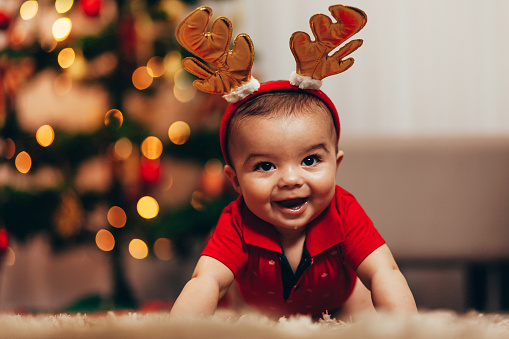 Cute baby boy wearing reindeer antlers crawling on floor over Christmas lights. Looking at camera. Holiday season.