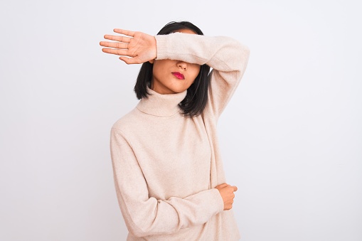 Young chinese woman wearing turtleneck sweater standing over isolated white background covering eyes with arm, looking serious and sad. Sightless, hiding and rejection concept