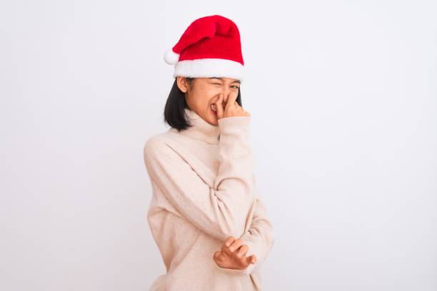 ung vacker kinesisk kvinna bär julen santa hatt över isolerad vit bakgrund luktar något stinkande och äckligt, outhärdlig lukt, hålla andan med fingrarna på näsan. dålig lukt koncept. - santa hat bildbanksfoton och bilder