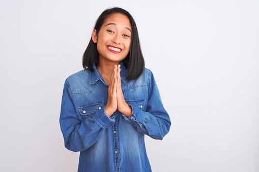 Young beautiful chinese woman wearing denim shirt standing over isolated white background praying with hands together asking for forgiveness smiling confident.