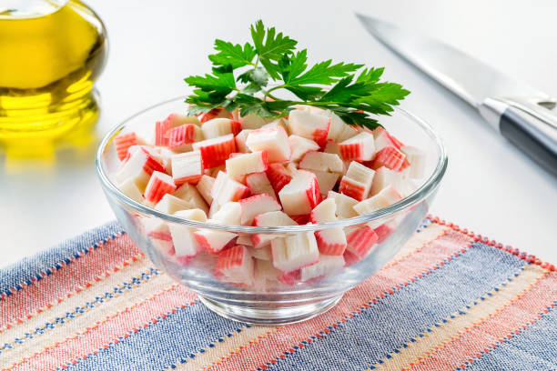Sliced surimi crab sticks in a glass bowl on a striped table mat near olive oil glass jar and chef knife. Seafood and ingredient for salads. Healthy eating and cook at home. stock photo