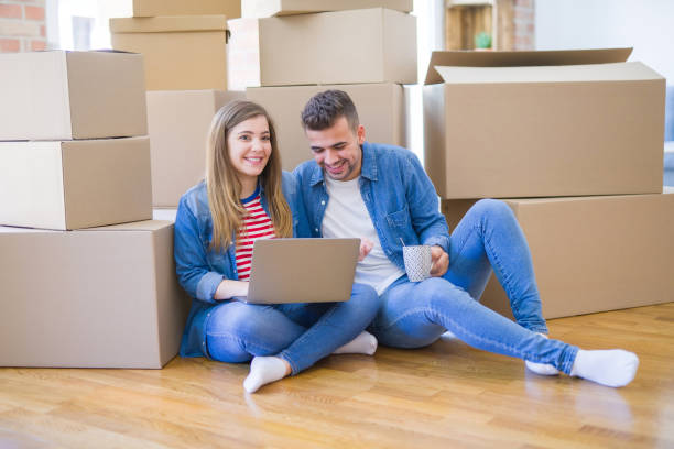 istock Young couple sitting on the floor around cardboard boxes using computer laptop, very happy moving to a new house 1185930143