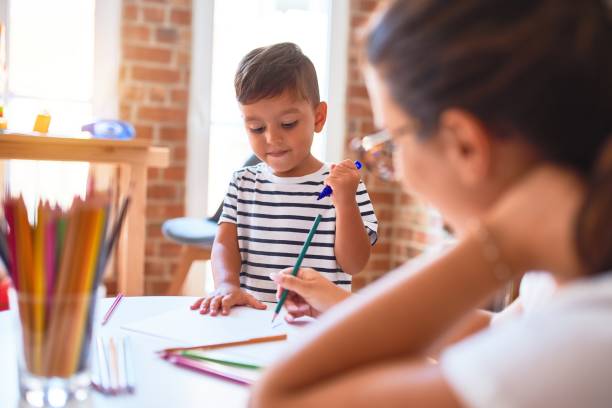 istock Beautiful teacher and toddler boy drawing draw using colored pencils at kindergarten 1185933175