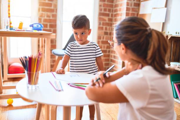 istock Beautiful teacher and toddler boy drawing draw using colored pencils at kindergarten 1185933184
